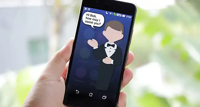 A closeup of a person's hand holding up a phone towards the camera. A clipart butler is on the screen and the text reads Hi Bob, how may I assist you?