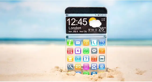 A thin transparent phone is propped up in the sand on a beach.