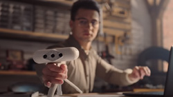 A person sitting at a desk is holding a white 3d scanner at arms length towards the camera. The capsule-shaped scanner is on a short tripod. The scanner has 3 small black circles in the front that are probably cameras or sensors.