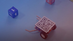 A closeup of a small three-wheeled square robot with a QR code on top. The robot has an open claw in front preparing to grasp a smaller numbered cube.