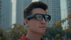 A closeup of a person wearing slightly bulky black AR glasses outdoors in the city.