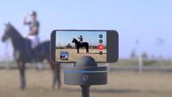 A view of a smartphone mounted horizontally on a dark gray camera mount that is on a tripod. The camera mount is hockey puck shaped with two semi-circle pieces coming out of the top that are holding the camera. A person on a horse can be seen in the blurred background and in focus on the smartphone.
