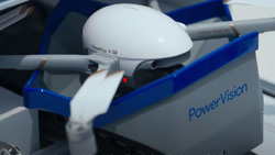 A white drone with a egg-shaped center has 4 struts leading from it with propellors on the end. It is sitting in a gray and blue docking station in a sliding drawer that is extended.