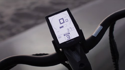 A closeup view from the rider's point of view of the black handlebars of an ebike. A smartphone sized monochrome LCD display with speedometer information sits in the middle.