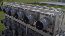 A partial view of a large machine in a grassy field during the day. It has a 6x3 array of fans and ducts that can be seen.