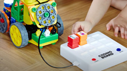 A closeup of a child's hands placing colored blocks on a white coding board. A wheeled robot is connected to the coding board via a cable.