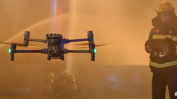 A drone hovers chest-high in front of a fireman in a smoky image.