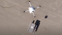 A view from above shows a drone lowering a package by cable to a park table