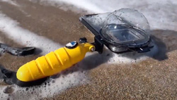 A smartphone is inside a clear, domed case with a yellow handle laying on the beach at water's edge