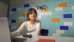 A person at a desk in front of a computer is looking at a small drone hovering above the desk