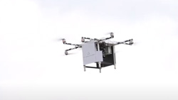A side view of a silver box-shaped quadcopter flying in overcast skies.