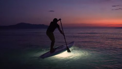 A view from the shore, shows a paddleboarder at dusk's end, standing and paddling over a small wave. There is a strong light underneath the paddleboard illuminating the ocean.