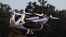 A black and white aircraft with four propellors facing upward is taking off just below the tree line. The cabin is shaped like a drop of water and has a large oval window in front. There are 2 fixed landing struts at the bottom