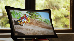 A rectangular display shows a dirt bike rider going around a corner. A window is behind showing bushes slightly out of focus.