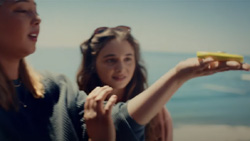 A view from their right-side, a person has their arm extended, palm up, holding a hand-sized yellow drone at the beach. A person looks on from behind.