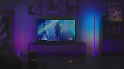 In a darkened room a television on a stand has a rock band displayed on the screen. Two purple and blue led lamps sit on each side. The lamps are minimilistic and look like a long pole rising from a cylinder base.