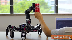 A black 6-legged robot with an extending arm is on a desk reaching for a can of soda being held up by a person.