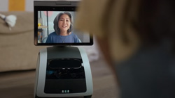 A view from behind a person's head shows a small gray and black device with a square base and a screen mounted on top. A person can be seen on the screen for a video chat.