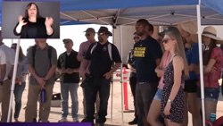 A group of 10 or more people in casual clothes are underneath a canopy perhaps in the desert. A person is hand-signing the audio in the upper left corner.