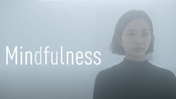 A woman is sitting meditating in a room full of mist. The word Mindfullness is written to the left in big letters