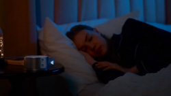 A night time scene shows a person sleeping on their side. A silver puck shaped device is on the nightstand in the foreground