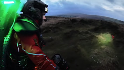 A closeup view from behind of a person in a red jumpsuit with a glowing green backpack on looking at the ground from above. A headlamp on their helmet is putting a spotlight on the rough terrain below.