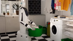 A white and black robot is carrying a green plastic laundry basket full of clothes to a white front loading washing machine