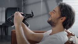 A view from the side shows a person sitting wearing a white tshirt in a tan leather chair holding a small black metal bare framed 4-legged robot. The person is making an expression like they are lovingly playing with a real dog