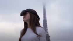 A person facing the camera is standing outdoors wearing a VR headset looking up and to the right. The Eiffel Tower is behind in foggy weather conditions.