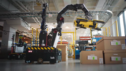 a wide view in a warehouse setting of a long-armed robot attached to a mobile base lifting Spot the quadruped robot