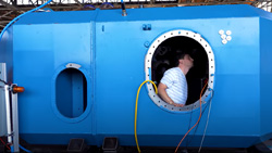 An ocean wave power device. A person is visible working inside a shipping container sized blue structure in drydock
