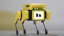 A closeup of a yellow and black quadruped robot. The face is a square white LCD screen with two almost square black eyes.