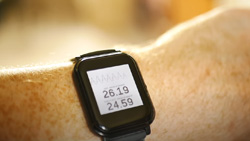 A closeup of a person wearing a black watch with a large display. The display shows a sine wave and the screen text reads 26.19 and 24.59