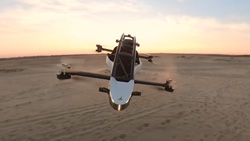 A view of a black and white, open framed VTOL from in front of the flying aircraft, at sunrise in the desert, with the horizon in the background. The craft is just wider than a person. It has 2 struts on each side, front and back, with 2 propellers, top and bottom, at the end of each strut.