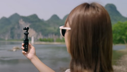 A closeup of a person facing a lake and mountain holding up a black toothbrush-sized camera