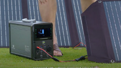 A gray rectangular box about the size of a desktop computer sits on grass. It has an LCD display in front. A power cord is plugged in the front. The device is surrounded by solar panels.