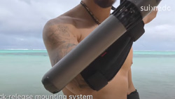 A closeup of a shirtless person on the beach holding a tube-like device on their arm up to the camera.