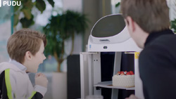 An adult and a child are looking at a robot that is delivering cake to their table. The robot is white and gray and has shelves underneath a LCD screen.