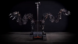A large all black robot with a treadmill platform base has two large arms on the sides with long 3-fingered claw grippers. The arms are attached to a large box near the base. A square beam with 2 cameras for eyes at the top extends upwards from the base.