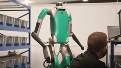 A green and silver humanoid robot is standing next to a person kneeling in front of a storage shelf. The robot has arms, legs, and a torso. The 'head' is a short silver cylinder with a black band around the middle