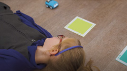 A child about 6 years old is lying on the floor on their back watching a robot car go over square, colored paper squares.