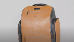 A closeup of a light brown and black backpack. It has a handle at the top and zippered pouches on both sides and the front. It has a small hole near the top for the camera