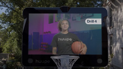 A view from the freethrow line shows a basketball backboard and hoop. The backboard is a display that shows a coach holding a basketball