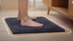 A closeup of a bare-footed person standing on a dark gray bathmat on a wood floor. An overlayed rounded white line surrounds the bathmat.