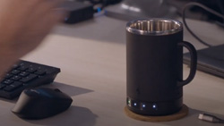 A closeup of a dark gray coffee mug with LED lights in the base is sitting on a desk with a mouse and keyboard.