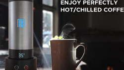 The Magic Brew coffee mug is tall, silver and black, and resting on a stand with the temperature showing. A steaming cup of coffee in a brown ceramic mug sits next to it.