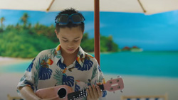 A young person is on a tropical beach under a white umbrella during daytime holding a ukelele and looking down at it. The person has a hawaiian shirt on and sunglasses on top of their head.
