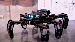 A closeup of a black metal six-legged open-frame robot. It has two glowing lights at the top front for eyes.