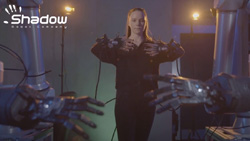 A person wearing an exoskeleton on their arms have their hands in front of them. In the foreground a pair of robot hands mimic the way the person is holding their hands.