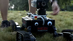A closeup view outdoors in a grassy field of a black rectangular robot with 2 plastic googly eyes in front. It has 4 jointed arms with tank-like treads at the end.